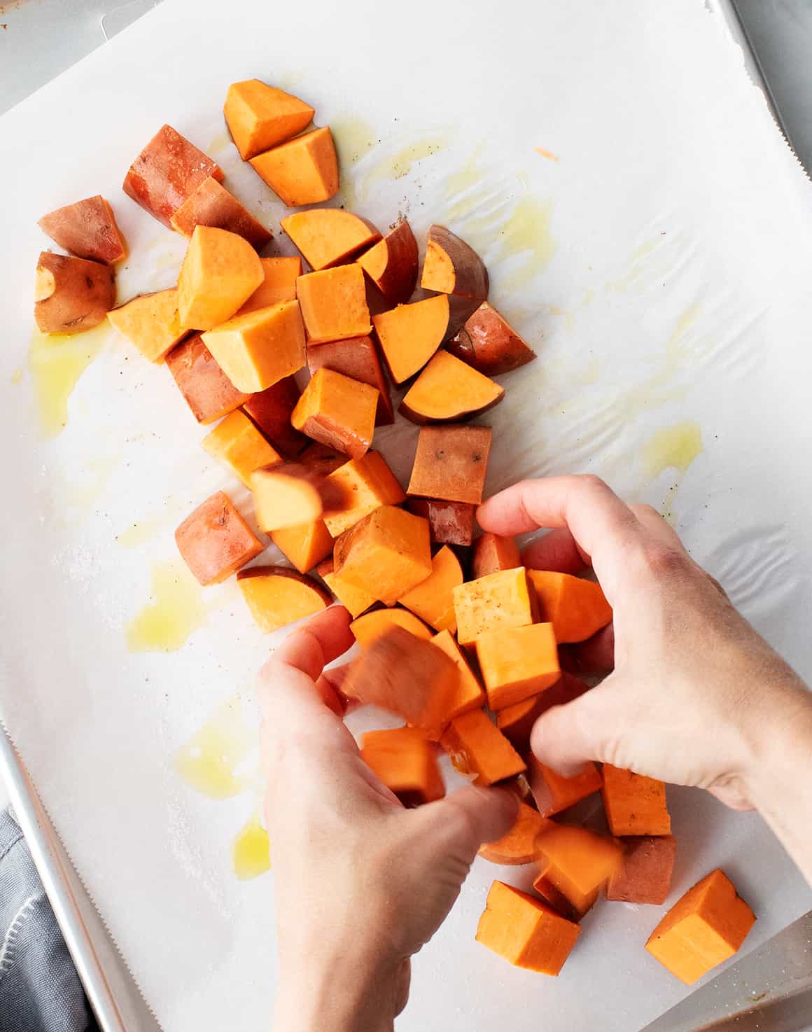 Hands tossing sweet potatoes with oil on a baking sheet