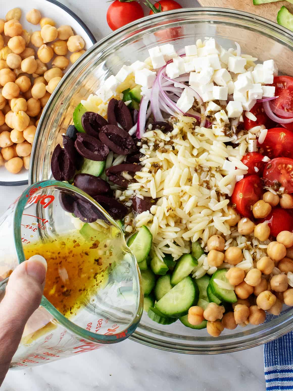 Hand pouring Greek dressing over bowl of pasta, olives, feta, and veggies