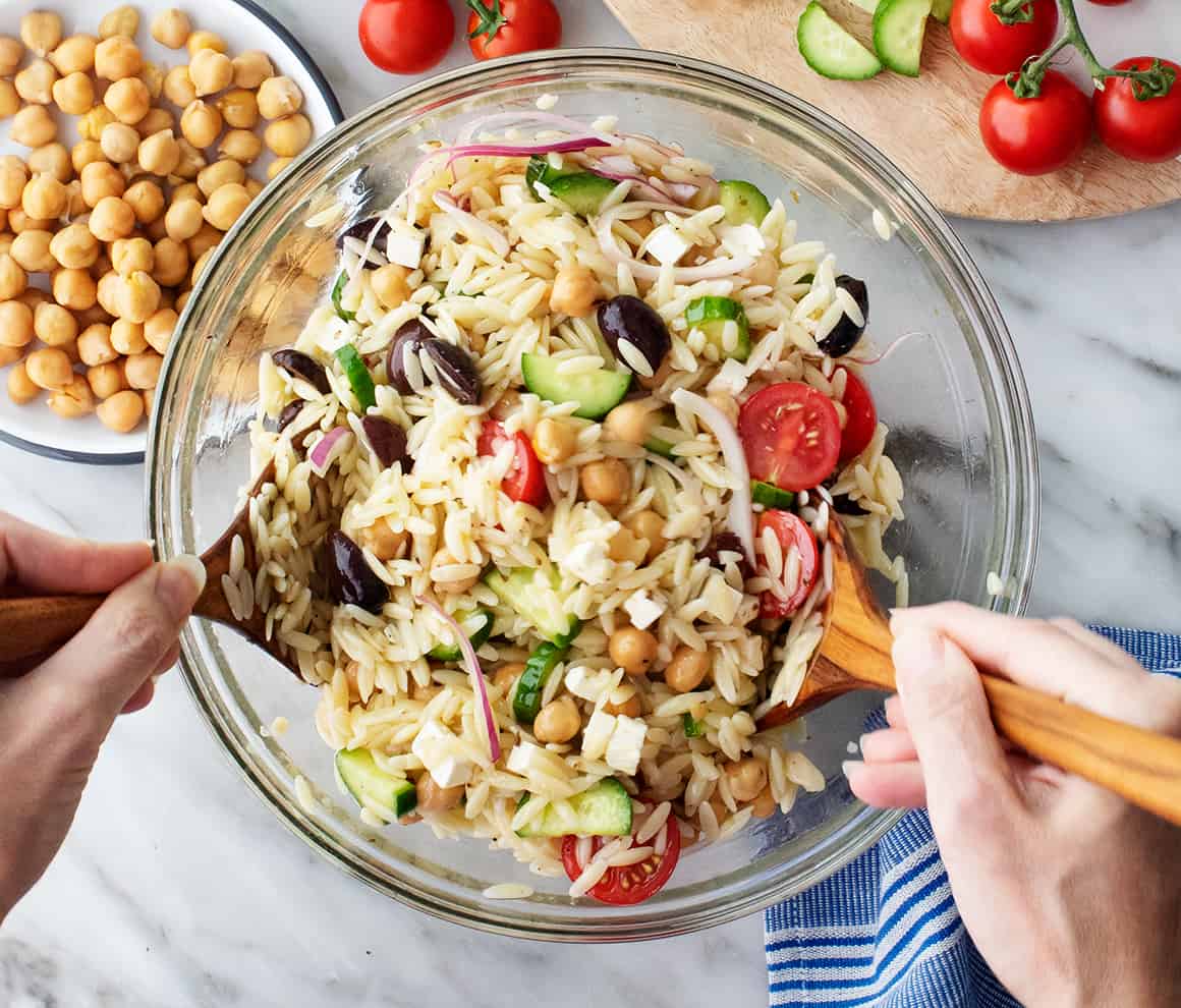 Hands tossing bowl of orzo pasta salad with wooden spoons