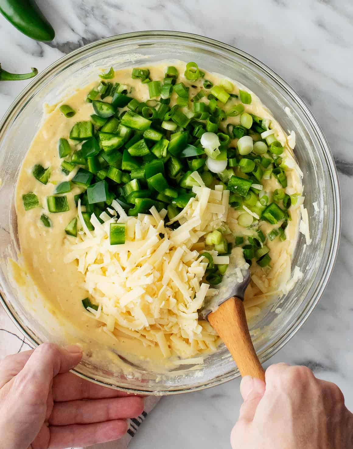 Hand folding jalapenos, cheddar, and scallions into bowl of cornbread batter with rubber spatula