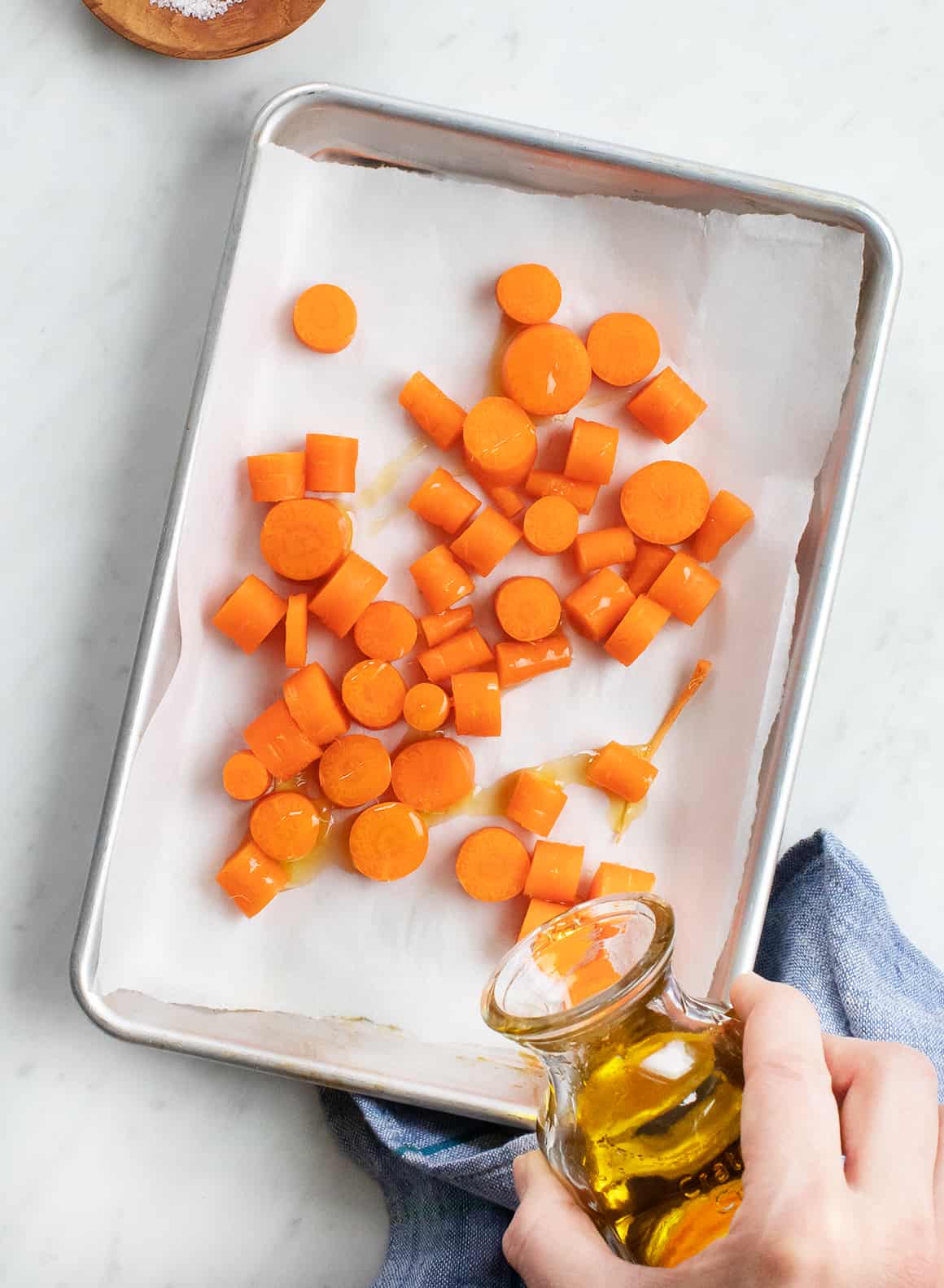 Hand drizzling olive oil over chopped carrots on a baking sheet