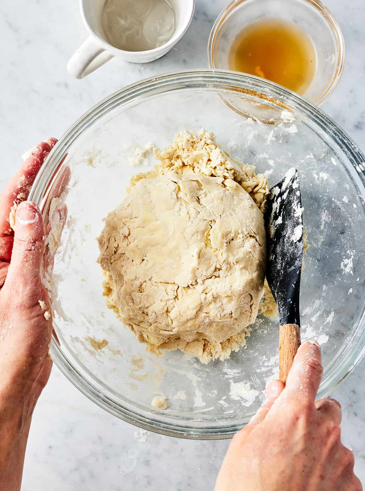 Hand using spatula to press homemade pie dough into a ball in mixing bowl