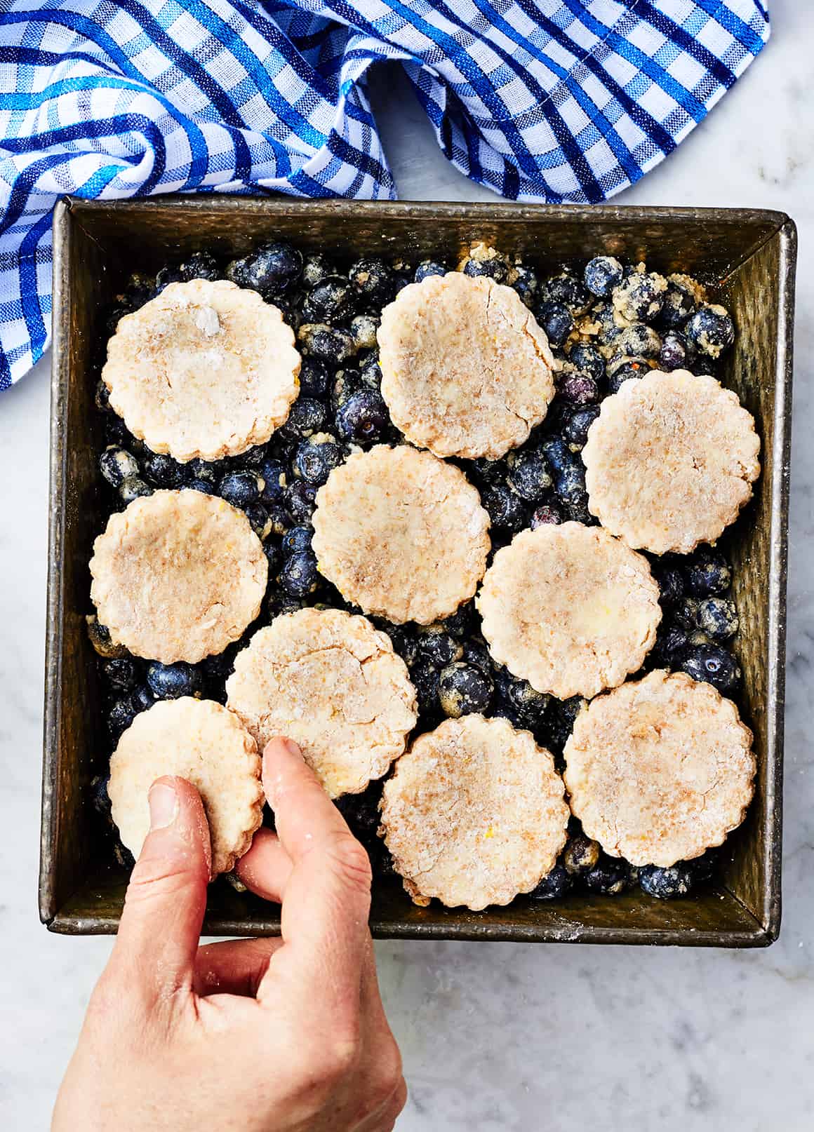 Hand placing final biscuit on blueberry cobbler in baking dish