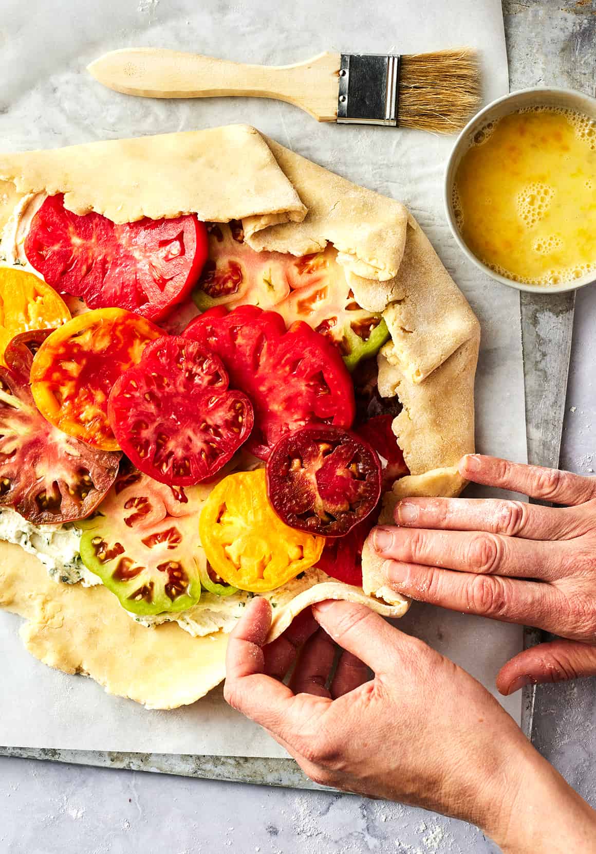 Hands folding edges of galette dough over sliced tomatoes