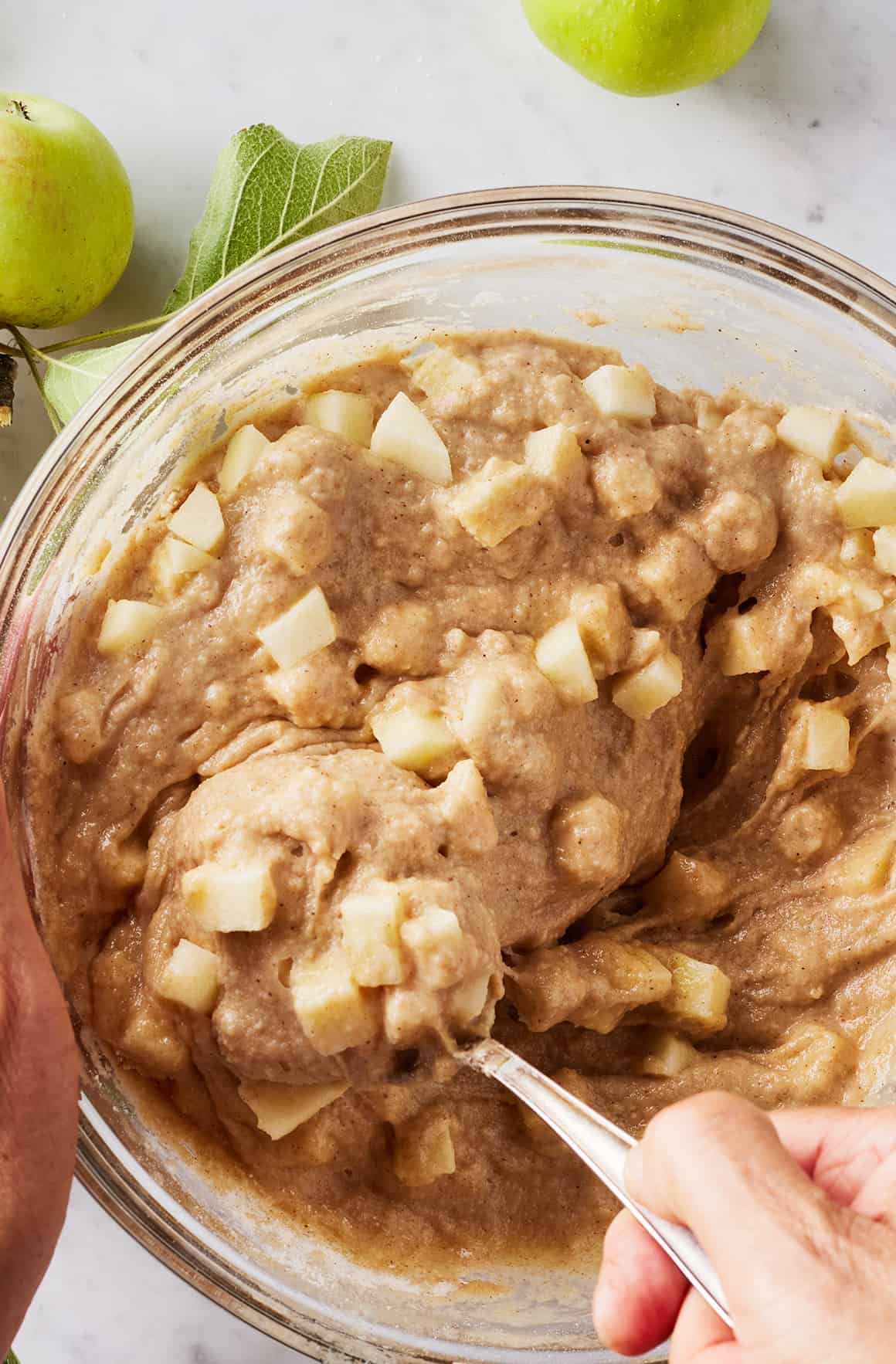 Hand using spoon to fold chopped apples into batter