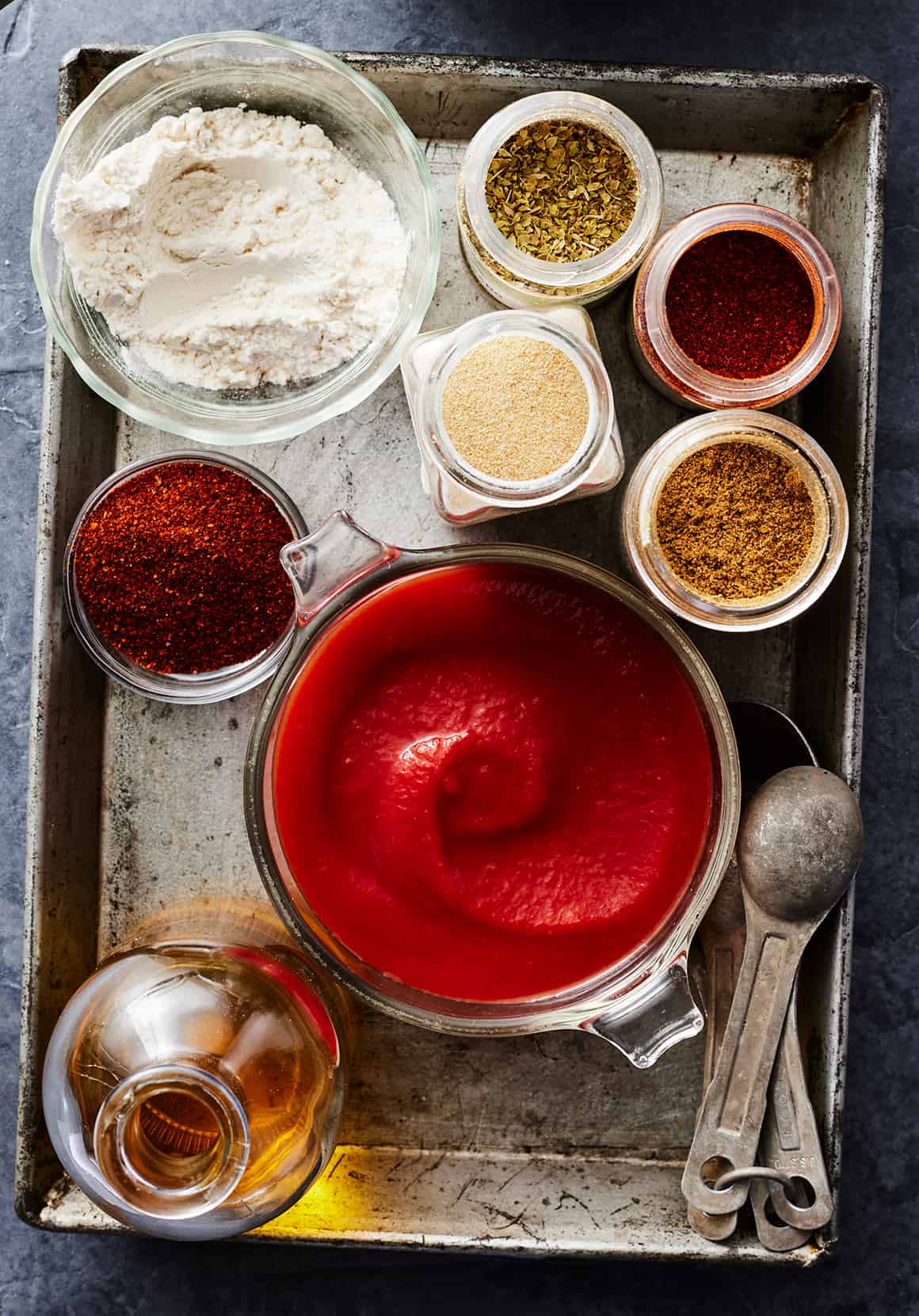 Overhead shot of tomato puree, avocado oil, spices, and all-purpose flour in small bowls on a metal tray