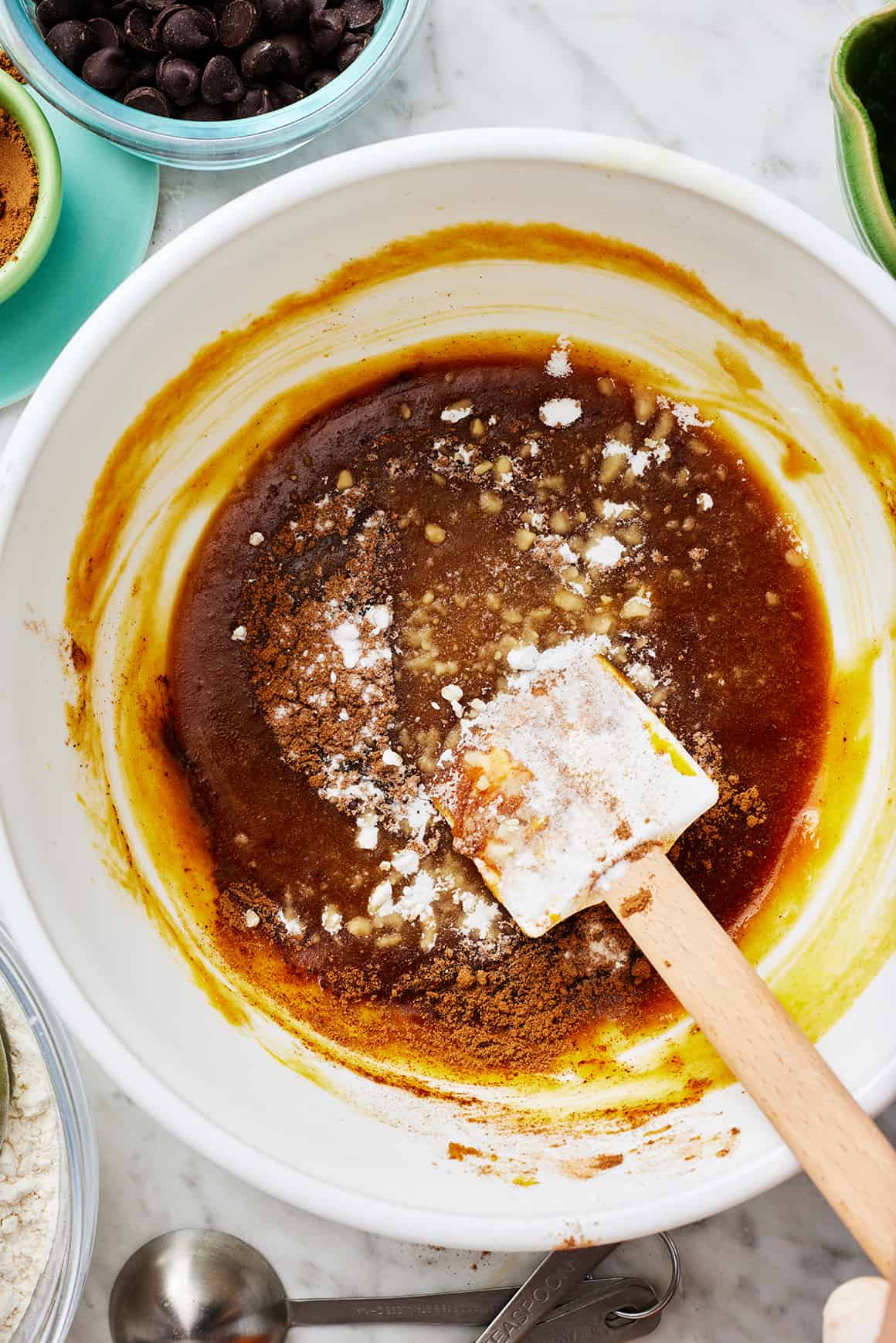 Dry ingredients sprinkled over wet ingredients in a mixing bowl with rubber spatula