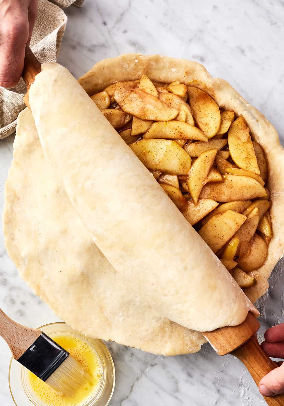 Hands using rolling pin to unroll top crust over apple pie