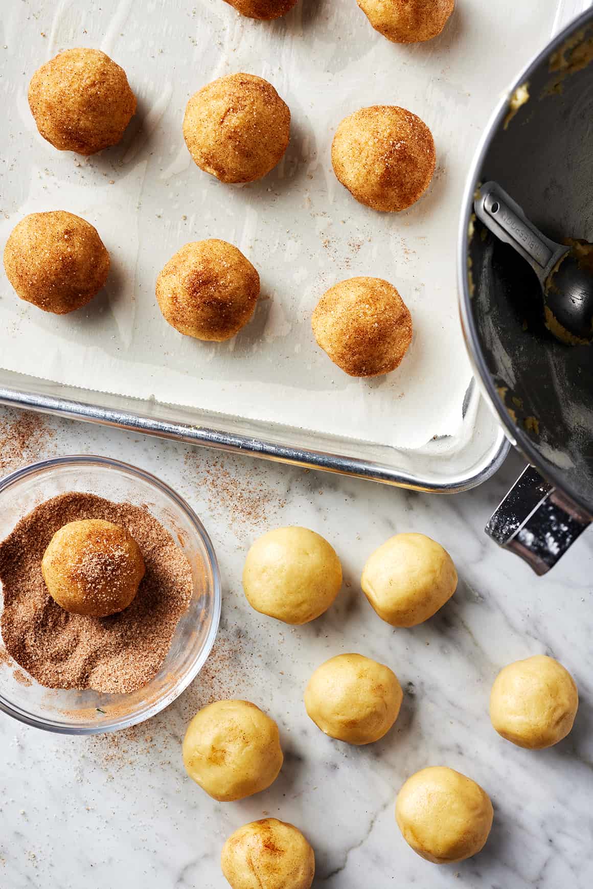 Balls of cookie dough on baking sheet next to small bowl of cinnamon sugar