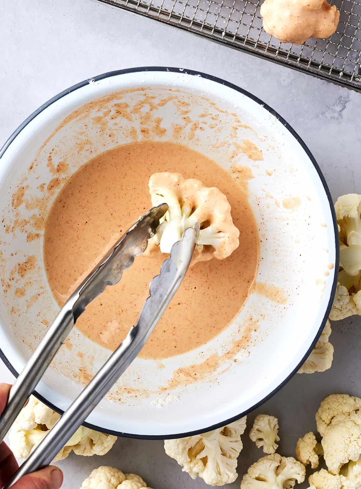 Hand using tongs to dip cauliflower floret in bowl of batter