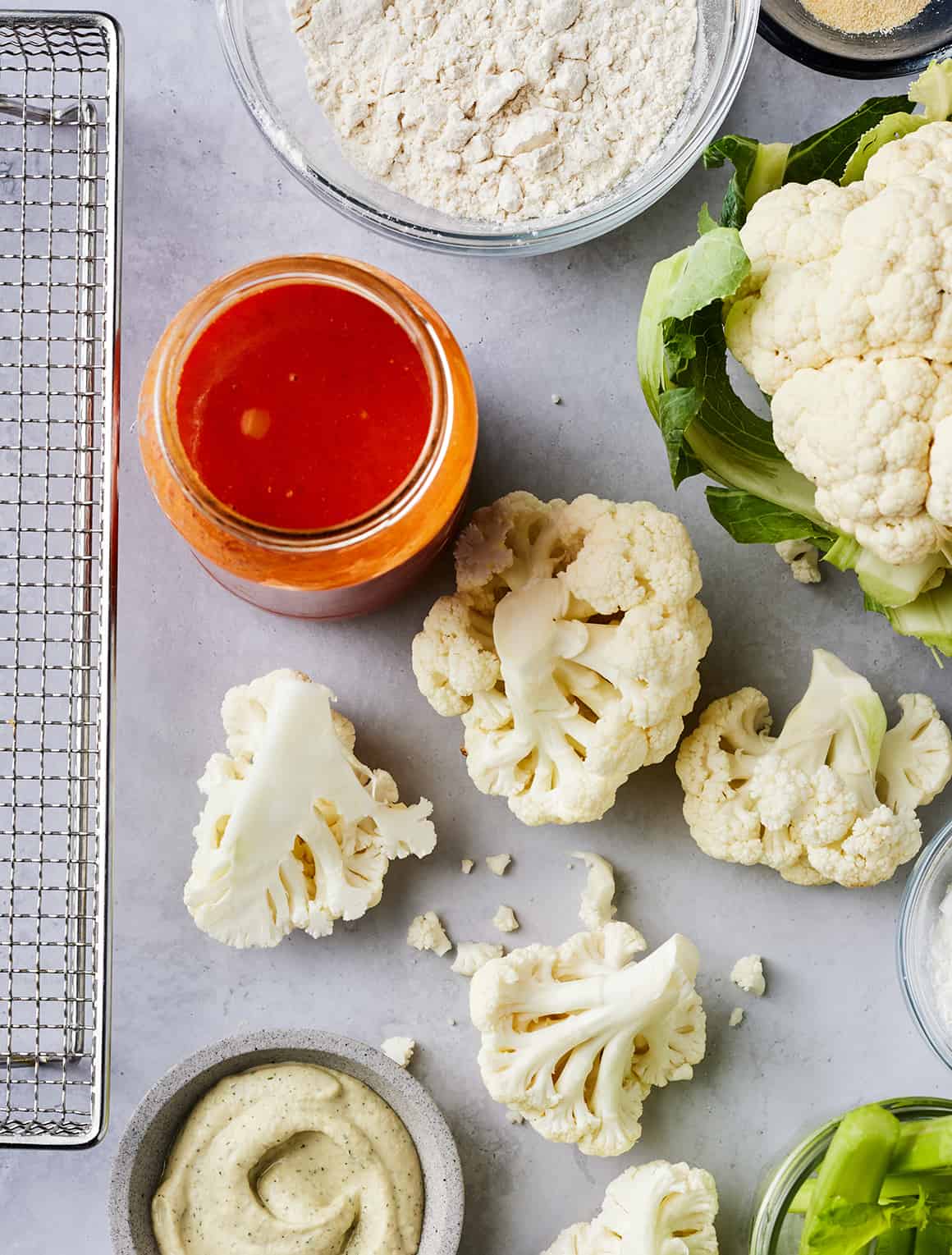 Cauliflower, jar of buffalo sauce, bowl of flour, and bowl of ranch dressing next to air fryer basket