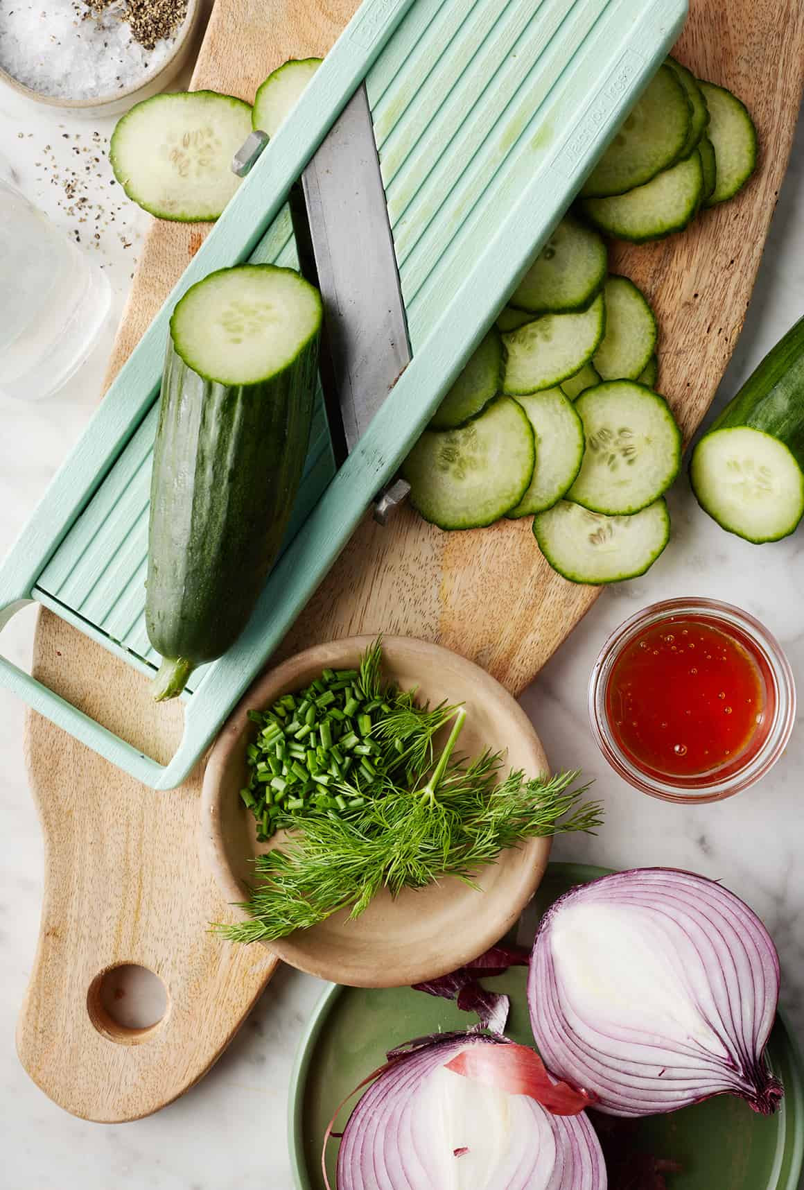 Thinly sliced cucumbers on cutting board next to red onion, dill, chives, and honey