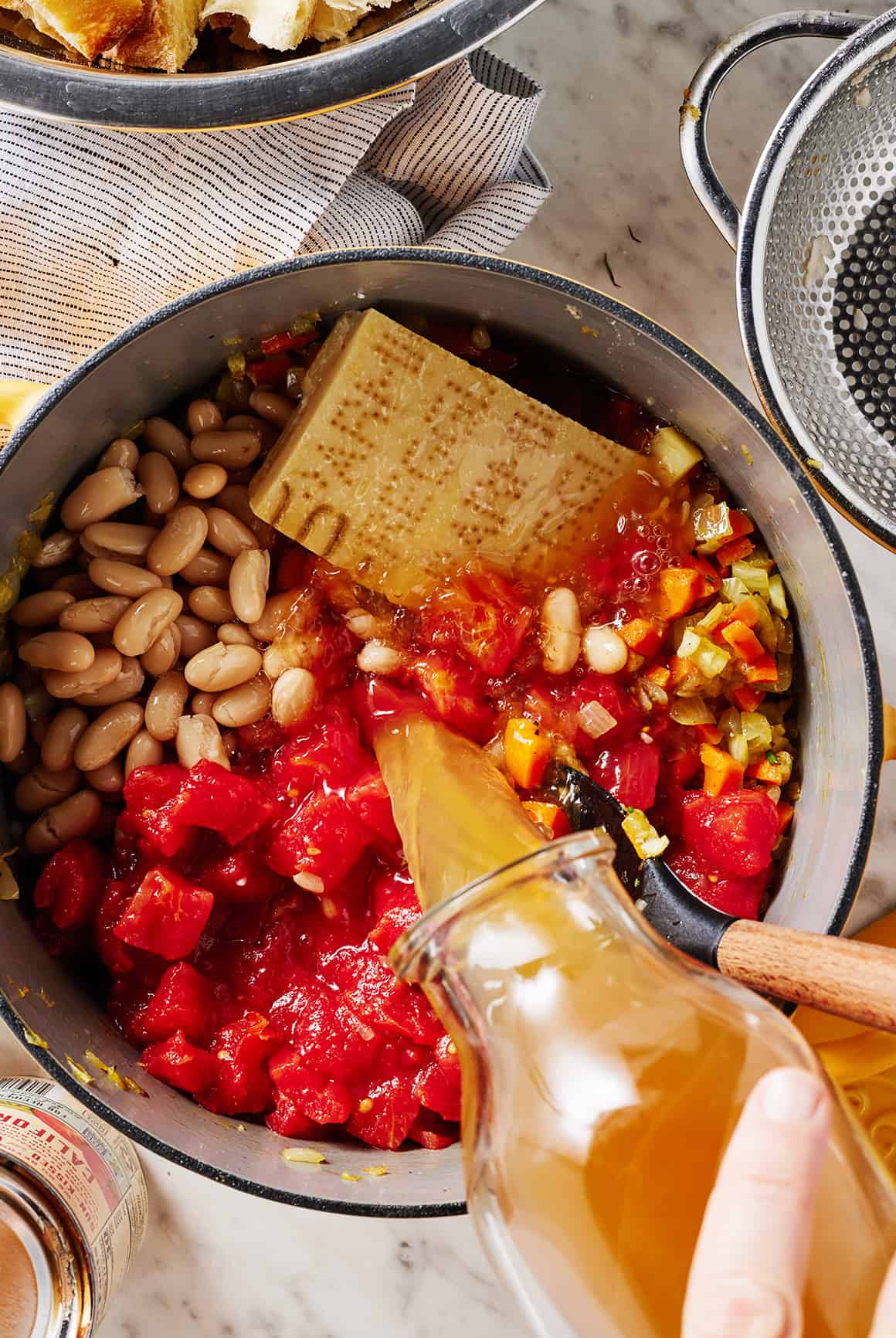 Pouring vegetable broth into soup pot with white beans, tomatoes, vegetables, and Parmesan rind