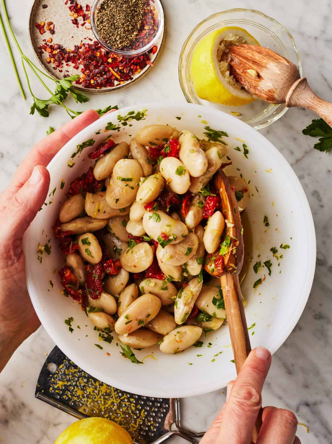 Stirring gigante beans, herbs, and sun-dried tomatoes in bowl with wooden spoon