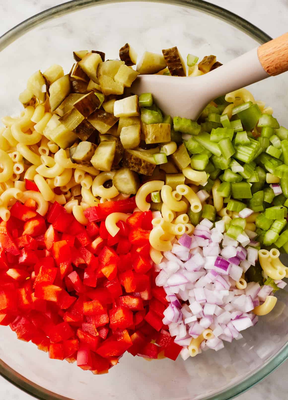 Chopped pickles, celery, red onion, red bell pepper, and pasta in glass mixing bowl