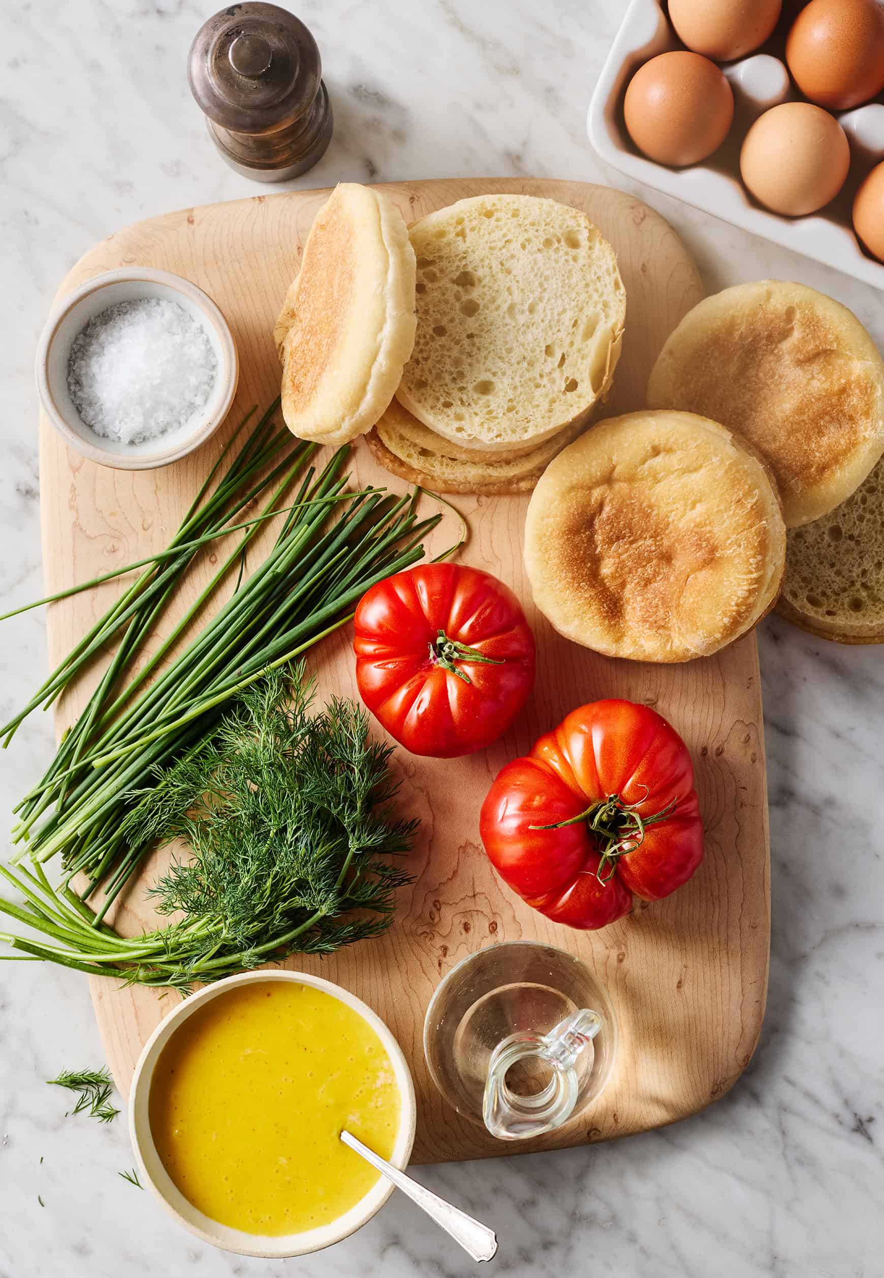 English muffins, hollandaise sauce, herbs, and tomatoes on cutting board next to eggs