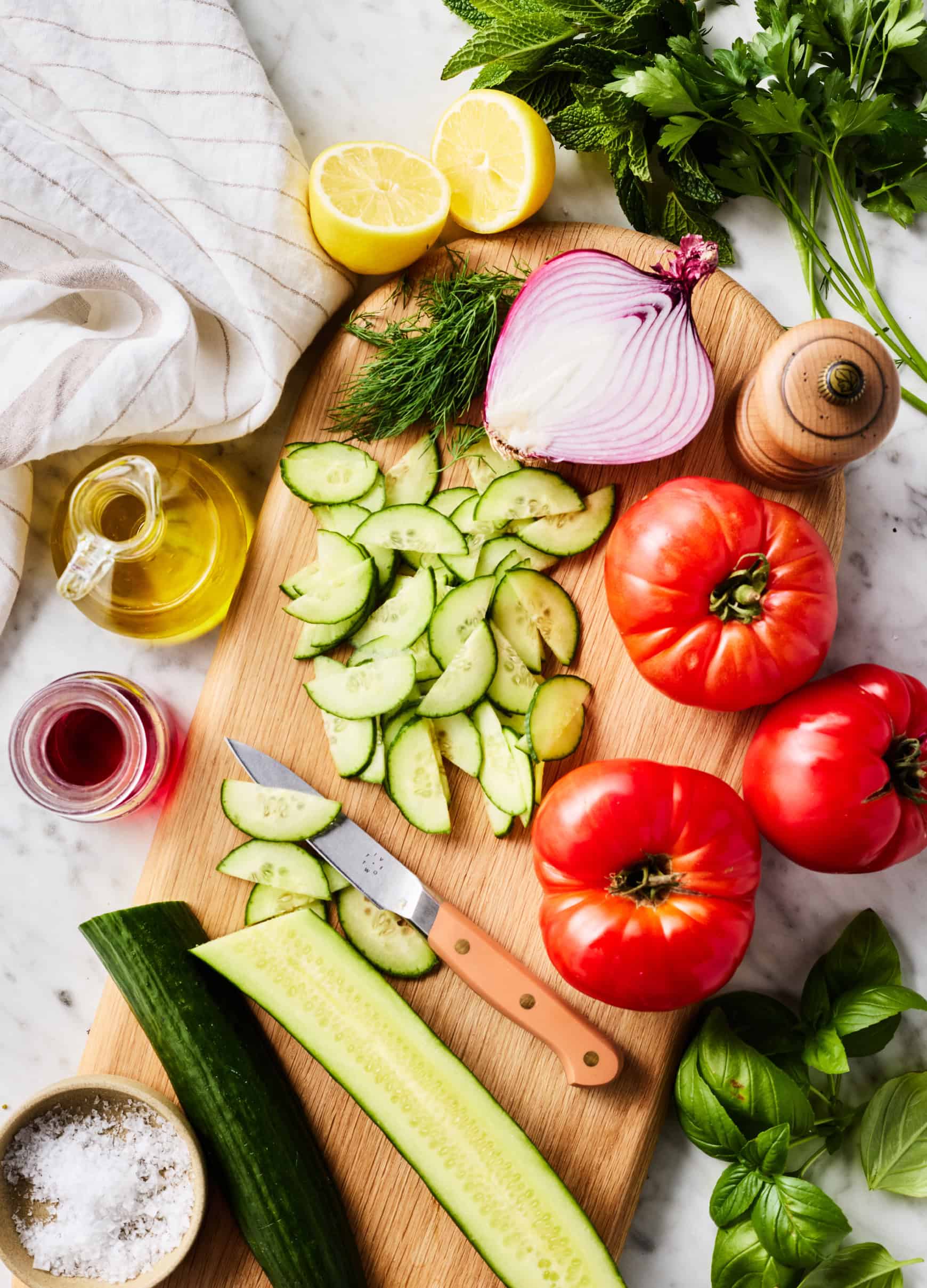 Sliced cucumbers, tomatoes, red onion, and fresh herbs on cutting board
