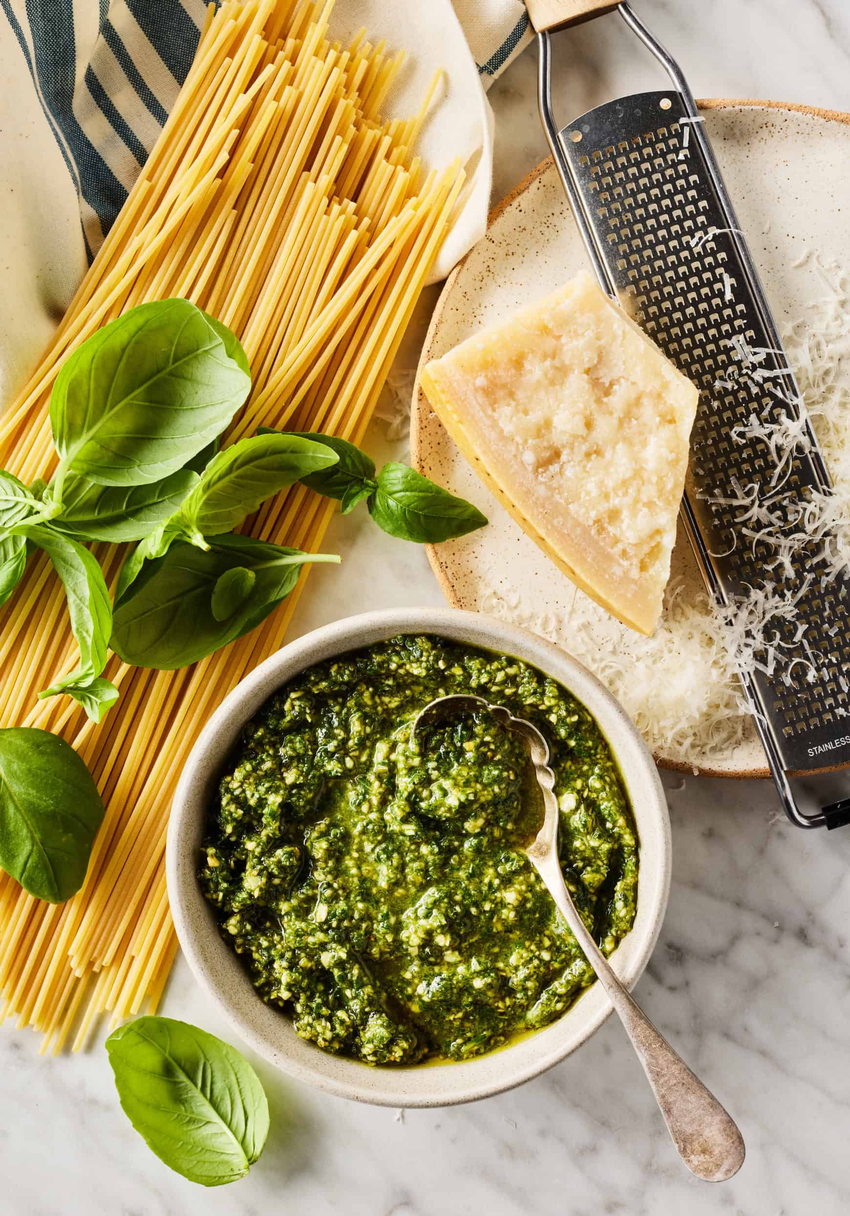 Basil pesto, fresh basil leaves, spaghetti, and Parmesan cheese on marble surface