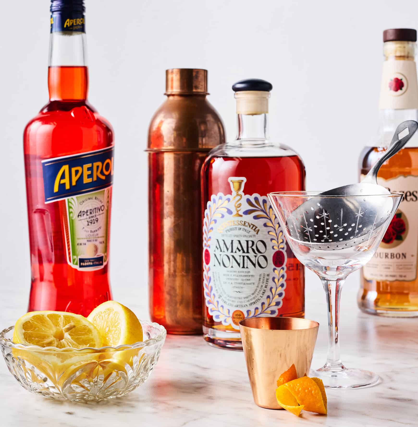 Recipe ingredients - Aperol, Amaro Nonino, bourbon, and fresh lemon - next to cocktail glass with orange twist