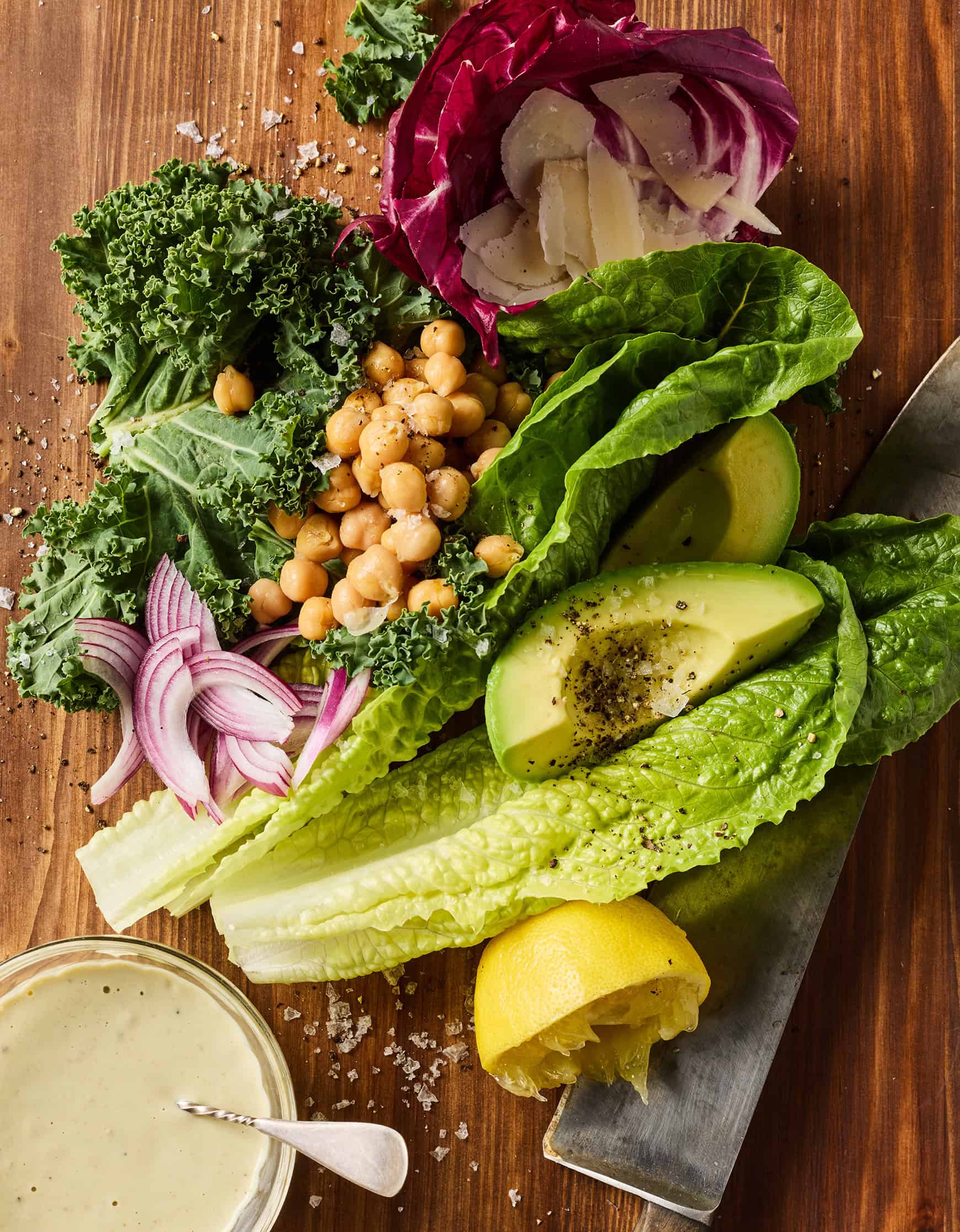 Romaine, kale, chickpeas, avocado, radicchio, onion, Parmesan, and lemon half on a cutting board with knife