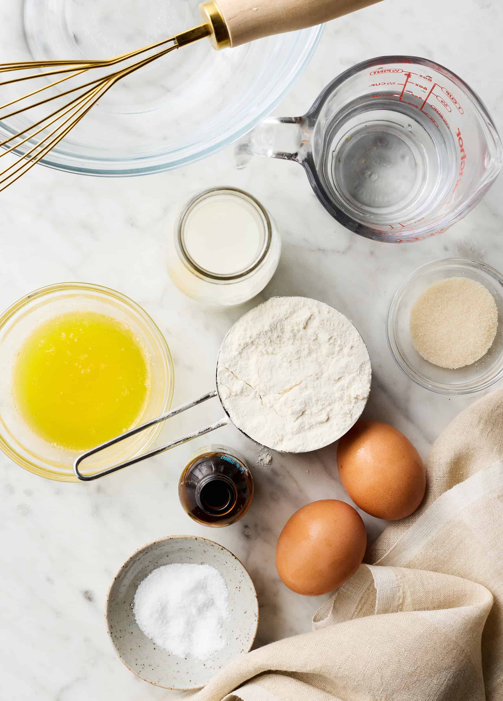 Crepe recipe ingredients on countertop - eggs, flour, milk, water, melted butter, sugar, vanilla, and salt