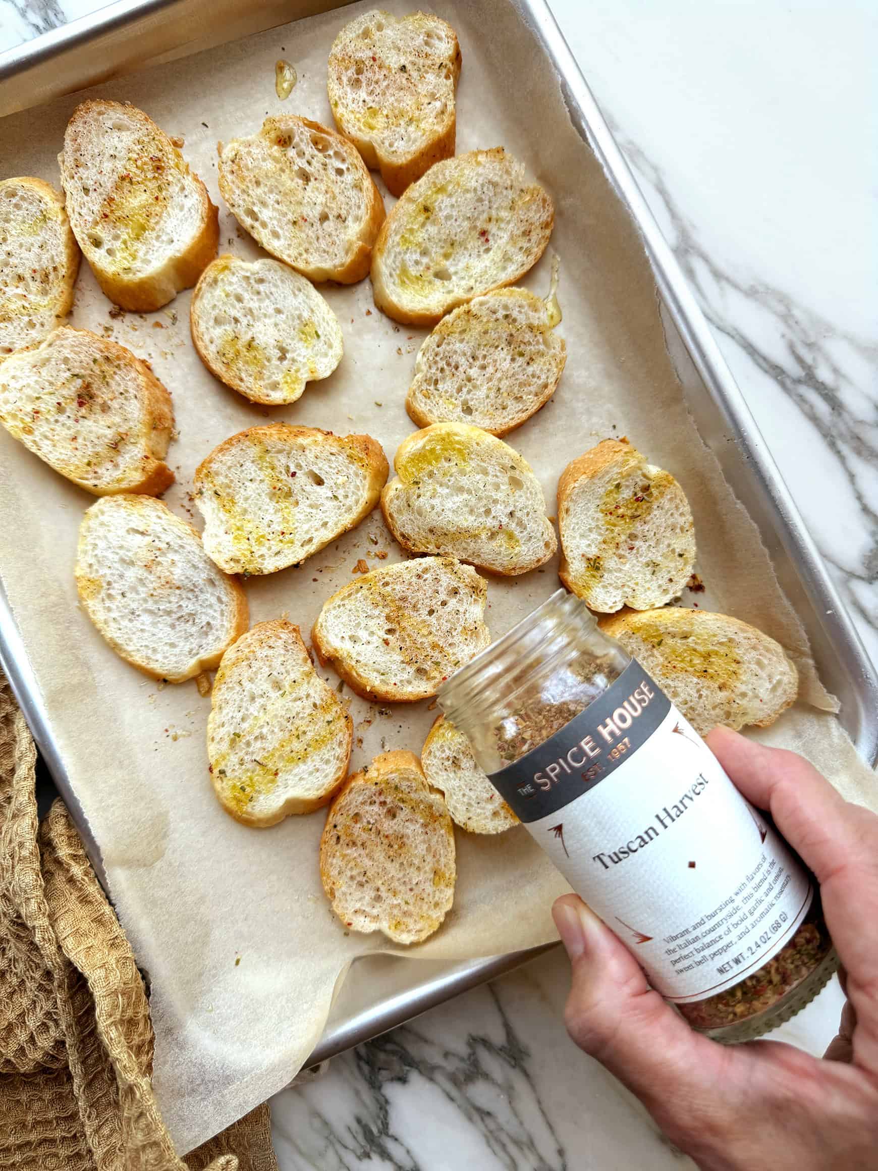 Sprinkling The Spice House Tuscan Harvest Seasoning over baguette slices on baking sheet