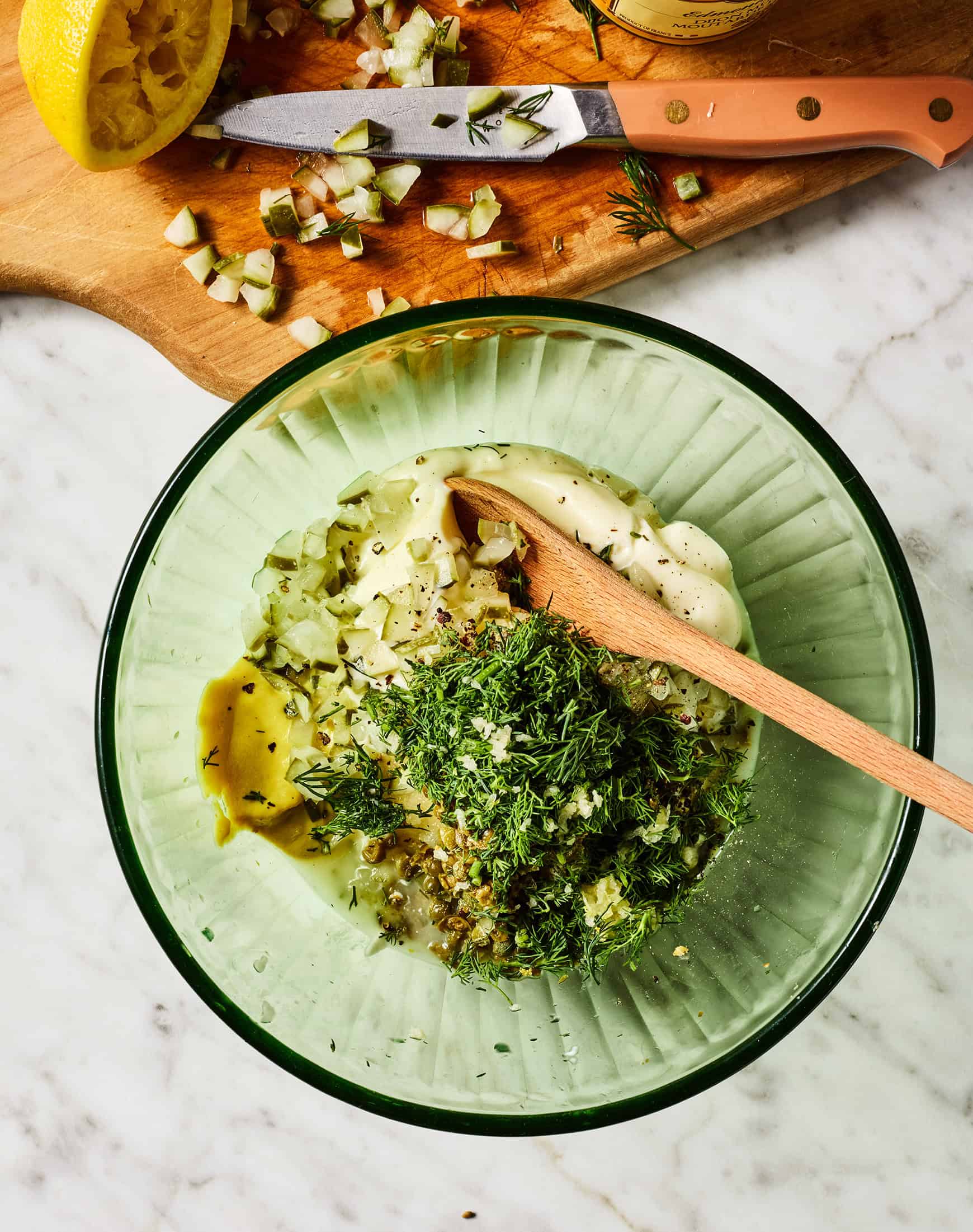 Mayonnaise, Dijon mustard, chopped pickles, capers, and fresh dill in mixing bowl with wooden spoon