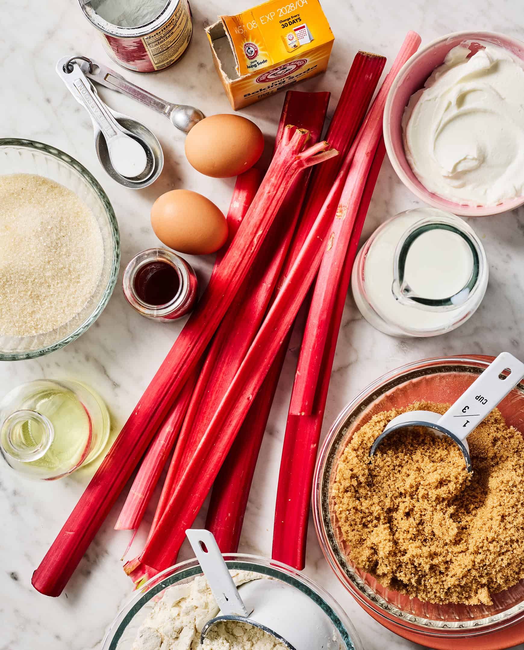 Recipe ingredients (fresh rhubarb, flour, brown sugar, cane sugar, milk, Greek yogurt, eggs, oil, vanilla, baking powder, and baking soda) on marble countertop