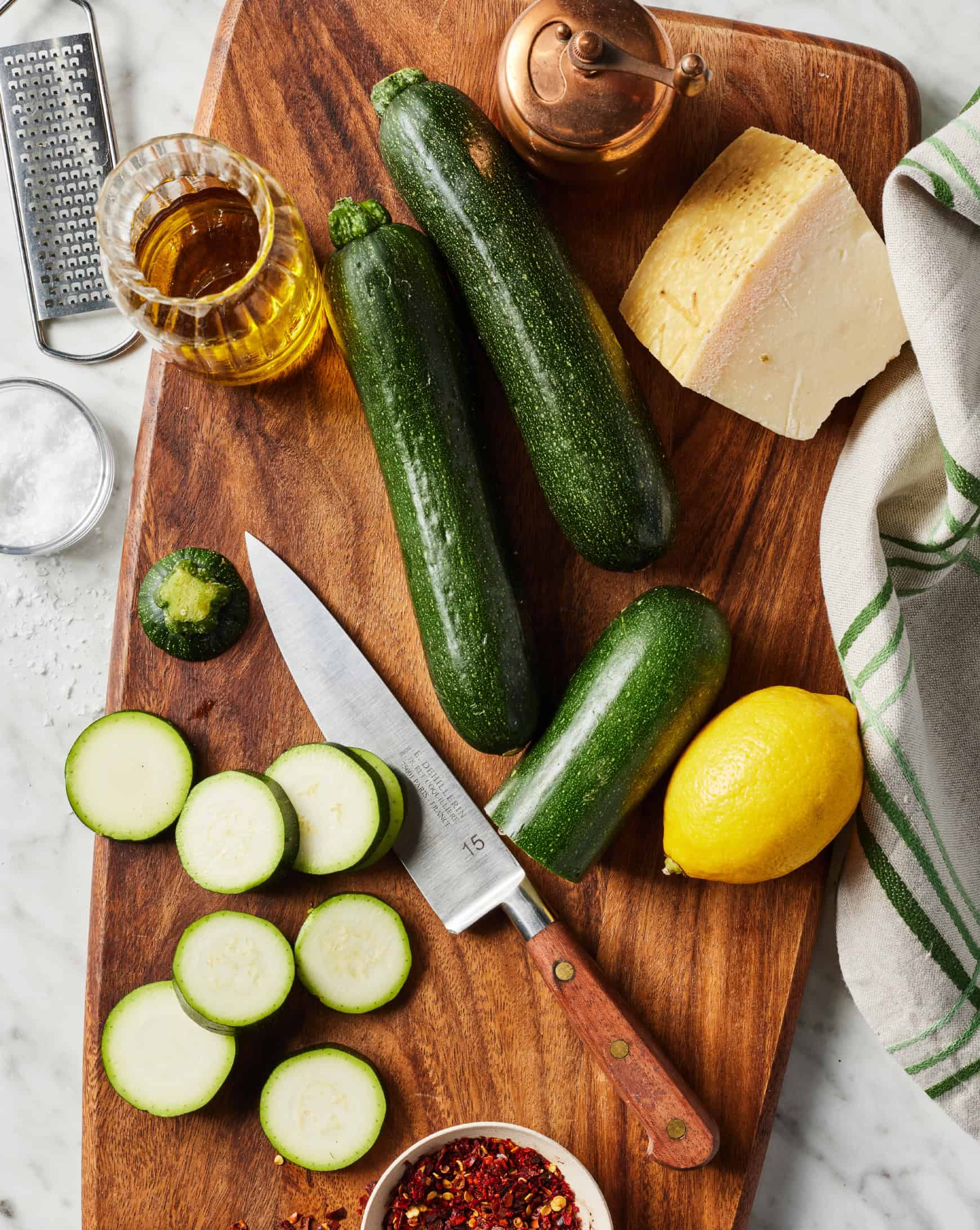 Two whole zucchini and one sliced zucchini on cutting board with lemon, red pepper flakes, olive oil, and Parmesan cheese