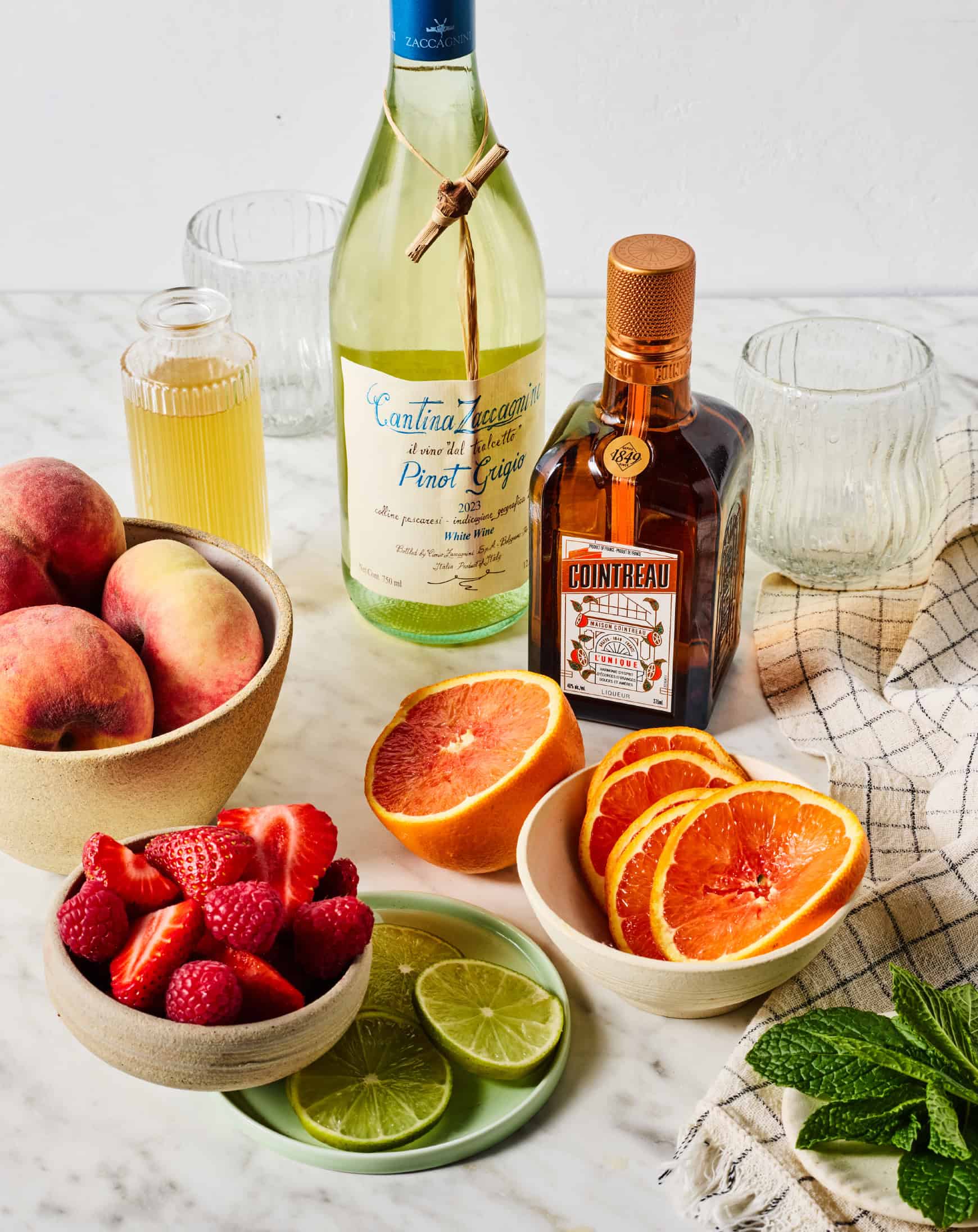 Recipe ingredients on countertop - white wine, Cointreau, simple syrup, oranges, peaches, berries, lime, and fresh mint