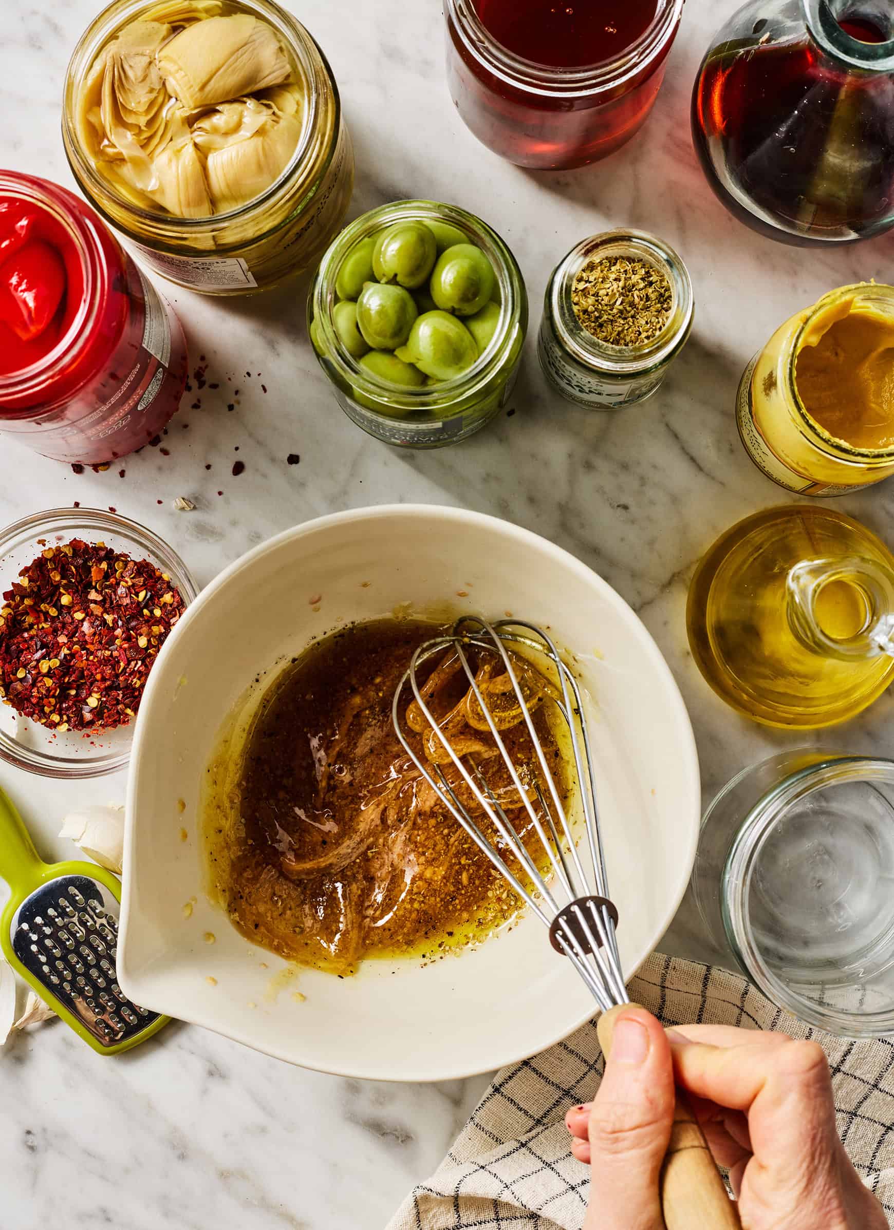 Whisking red wine vinaigrette dressing in medium bowl next to jars of olives, roasted red peppers, and artichoke hearts
