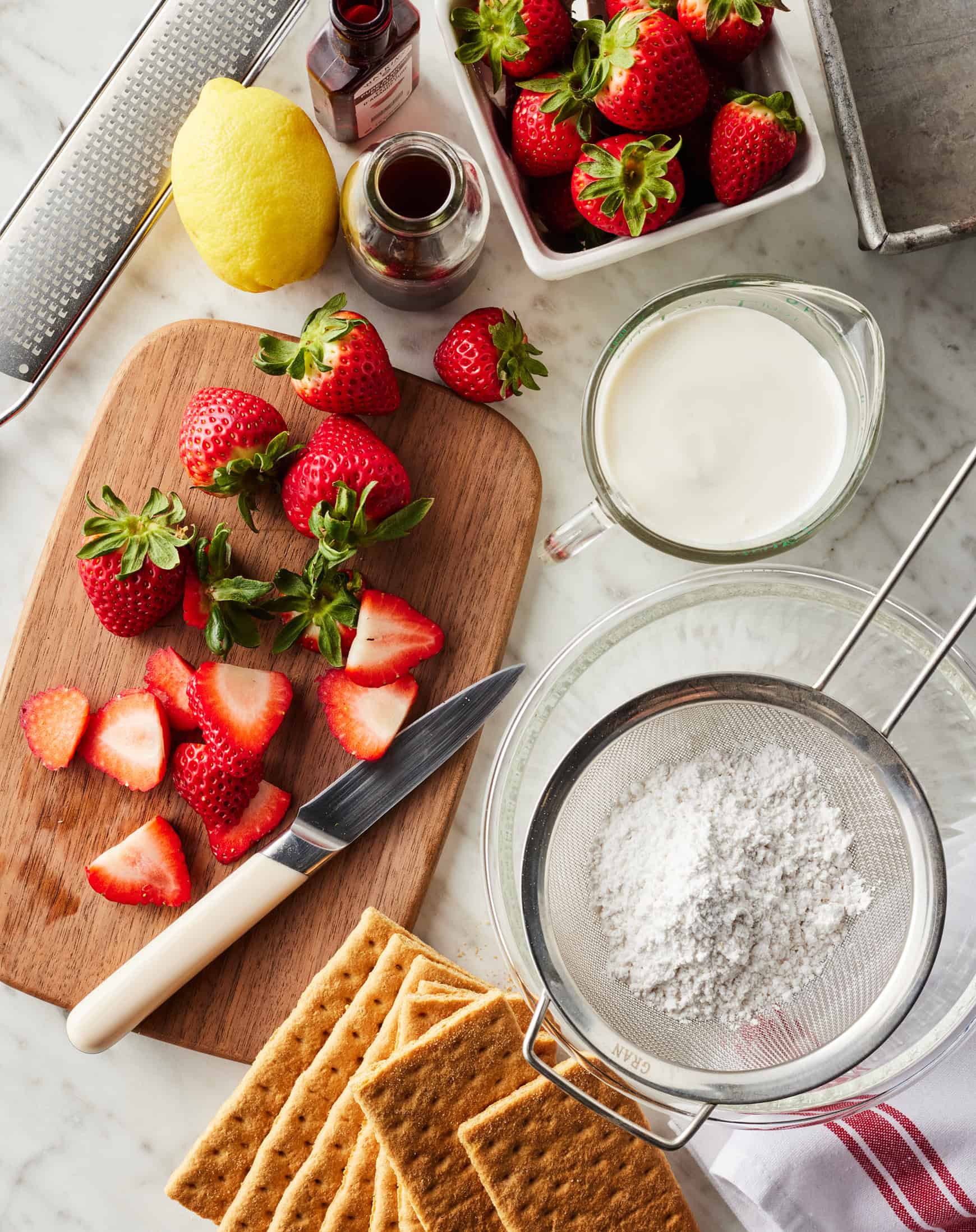 Sliced strawberries, graham crackers, cream, powdered sugar, lemon, vanilla extract, and almond extract on marble countertop