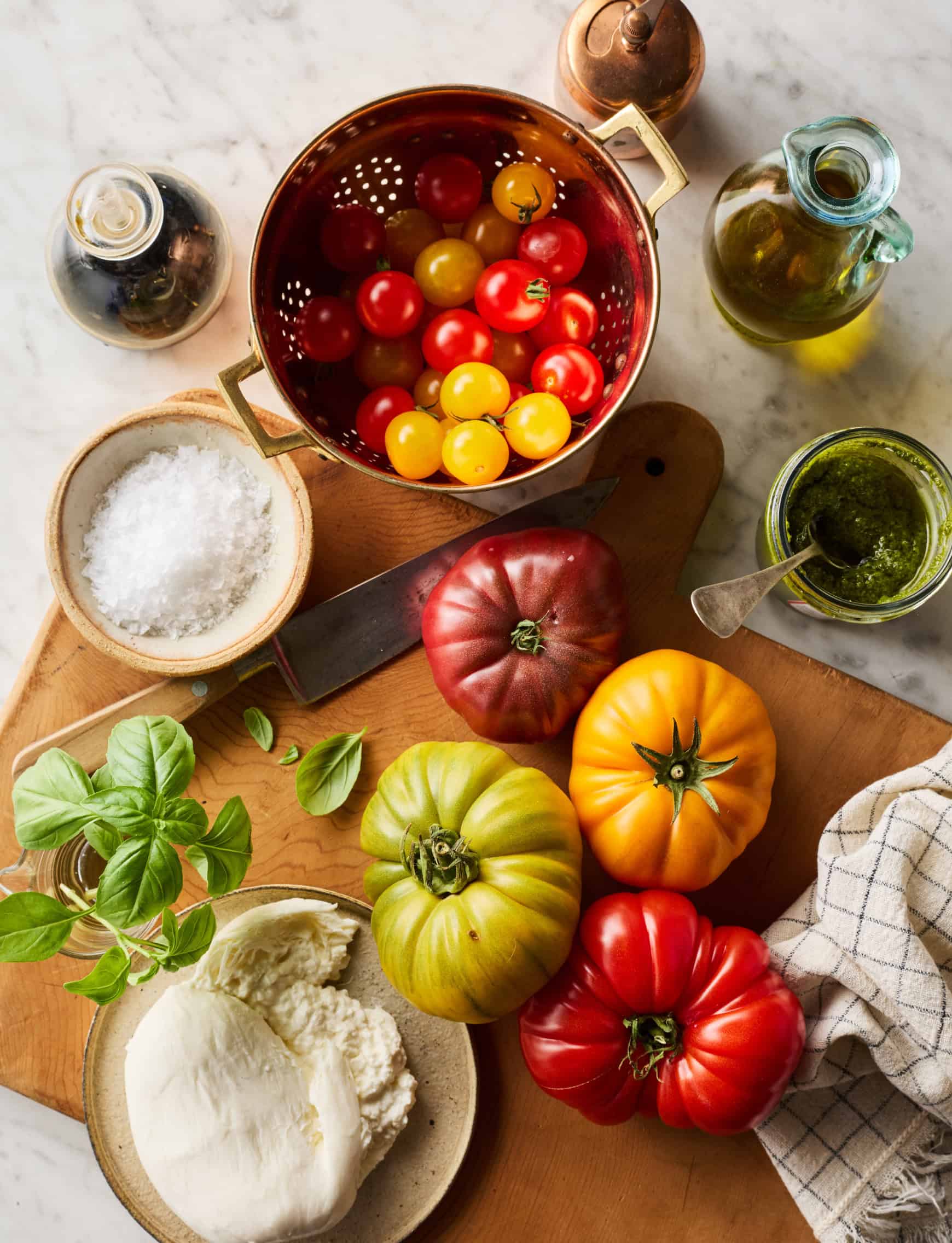 Burrata salad ingredients on marble countertop - tomatoes, burrata cheese, basil, pesto, flaky salt, olive oil, and balsamic