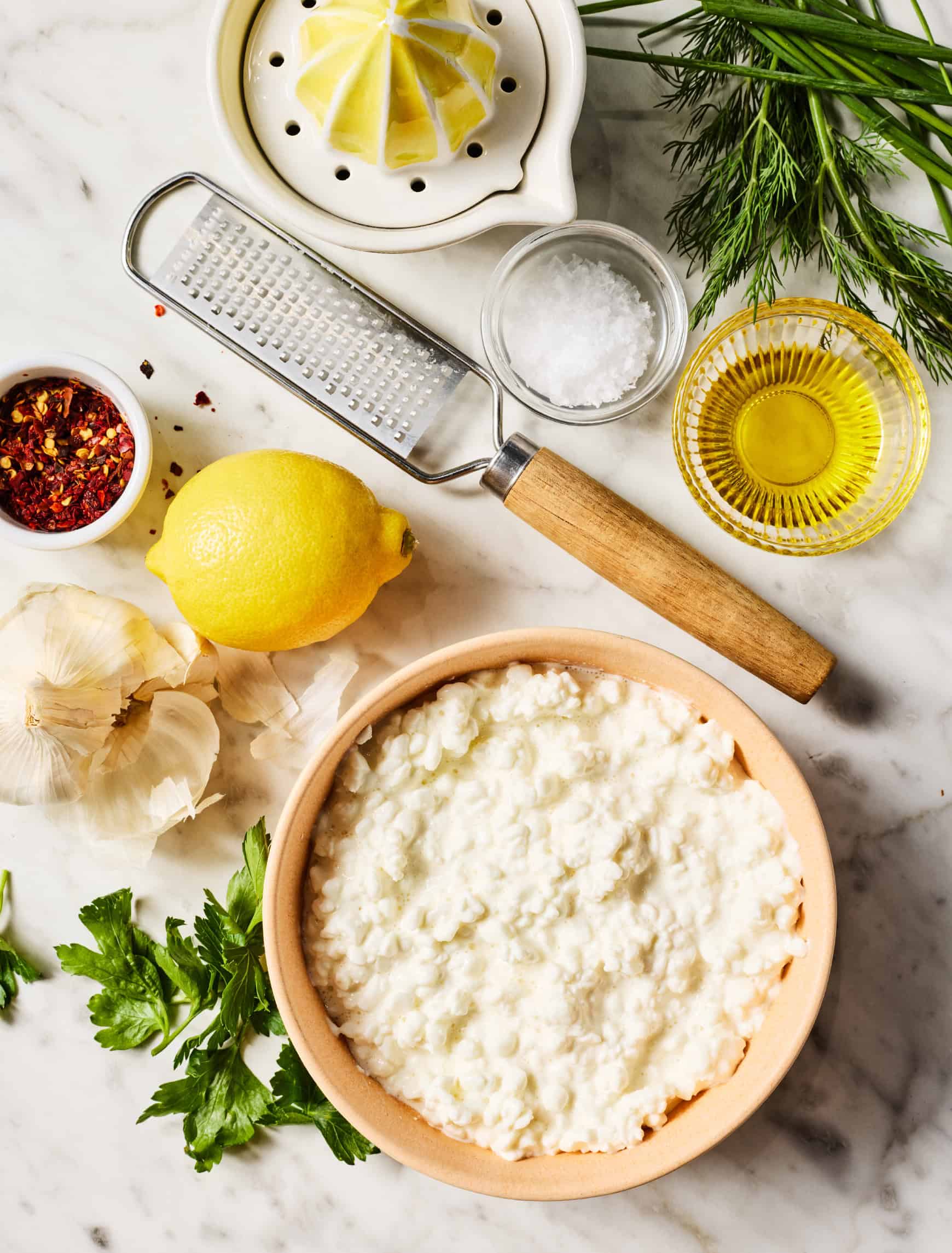 bowl of cottage cheese on marble countertop next to olive oil, lemon, salt, herbs, garlic, and red pepper flakes