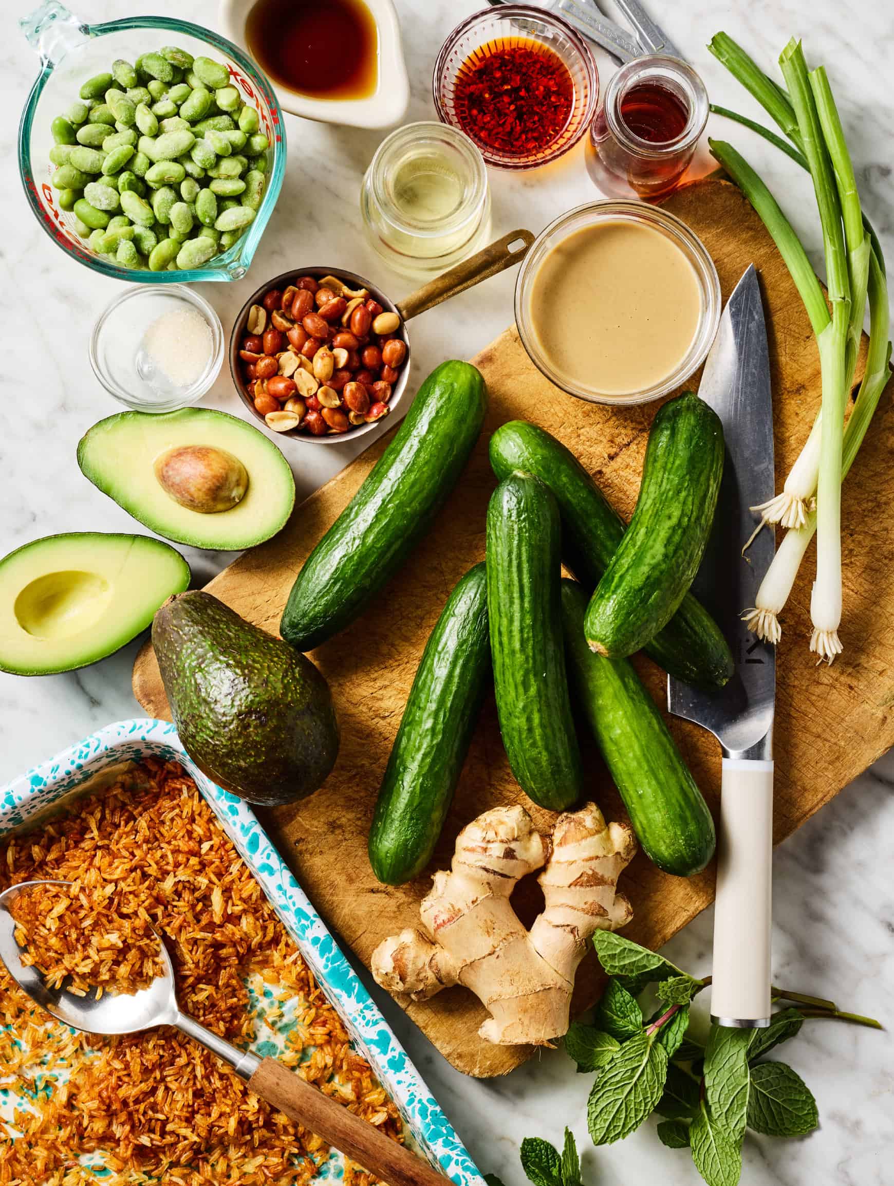 Recipe ingredients on countertop - crispy rice, cucumbers, avocado, peanuts, edamame, green onions, mint, ginger, tahini, rice vinegar, maple syrup, tamari, and chili crisp