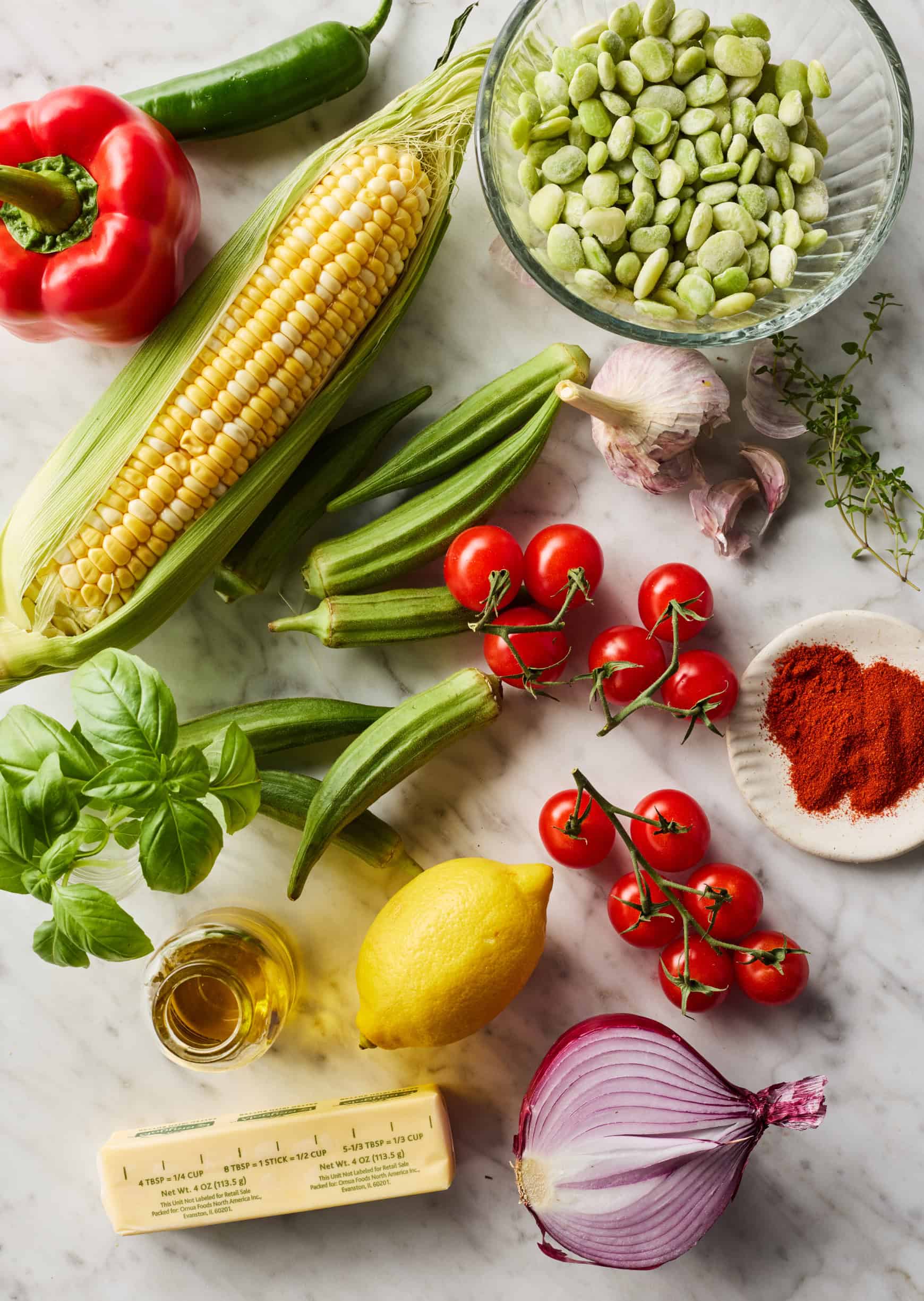 Recipe ingredients on countertop - fresh corn, peppers, cherry tomatoes, okra, frozen lima beans, smoked paprika, garlic, red onion, olive oil, butter, and herbs