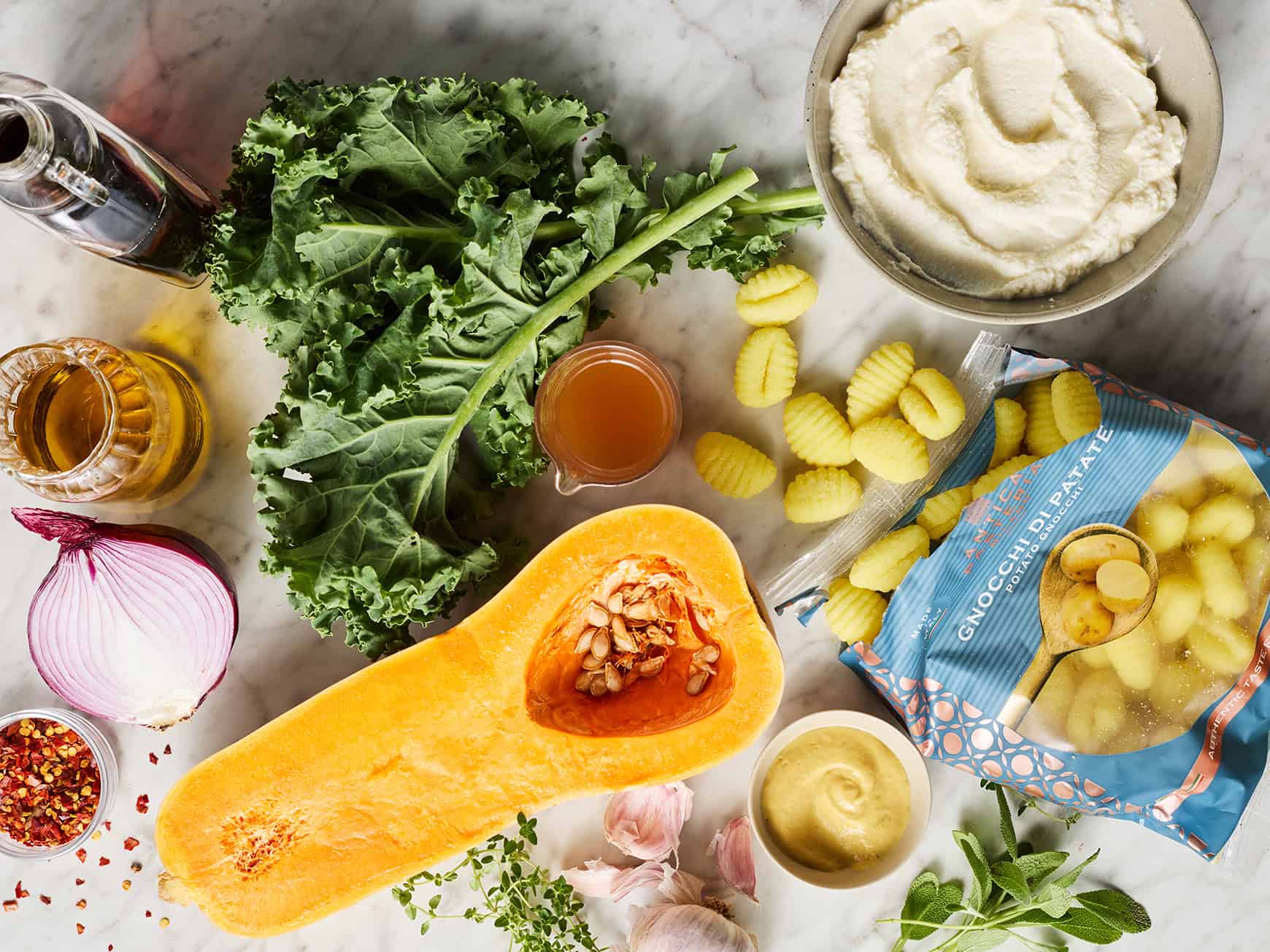 Recipe ingredients on countertop: butternut squash, kale, red onion, ricotta, gnocchi, herbs, Dijon mustard, apple cider vinegar, and olive oil